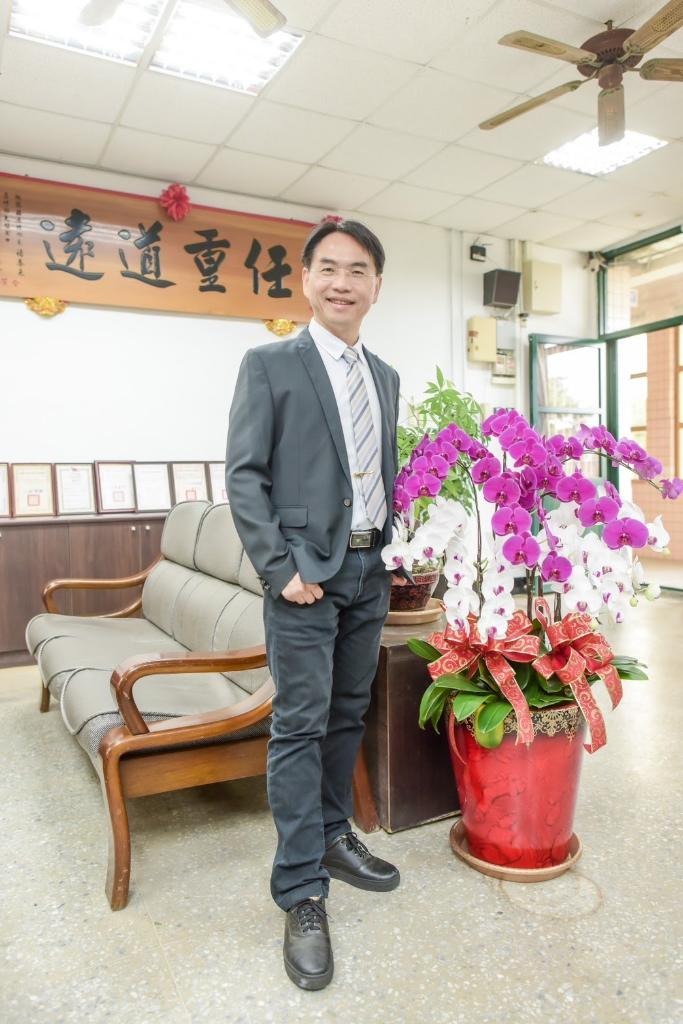 觀音國中黃博欽校長於校長室與蘭花盆栽合影,背景有「遠道重任」匾額